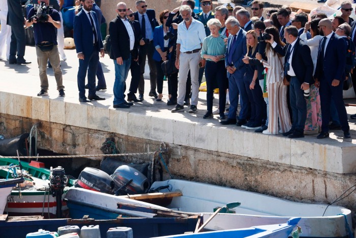 A group of well dressed people stand on a dock beside rowing boats