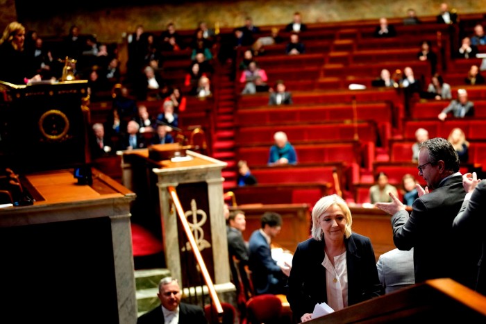 A woman walks away from a podium in a large auditorium with red seats and sprinkled with audience members