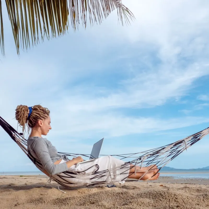 woman working on laptop lying in hammock at sand beach 