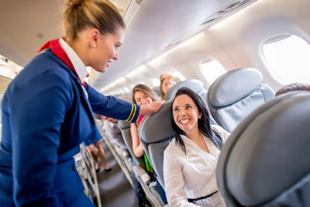 Woman sitting in airplane cabin smiling at cabin crew member — Getty Images