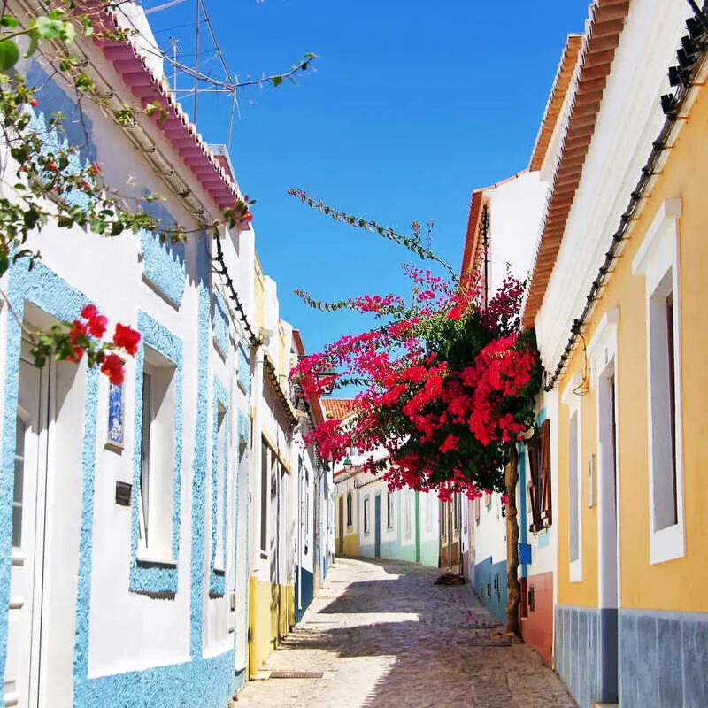 Whitewashed Houses With Colorful Door Frames And An Overhanging Bougainvillea Arching Over The Cobbled Street In Ferragudo, Algarve, Southern Portugal, Southern Europe