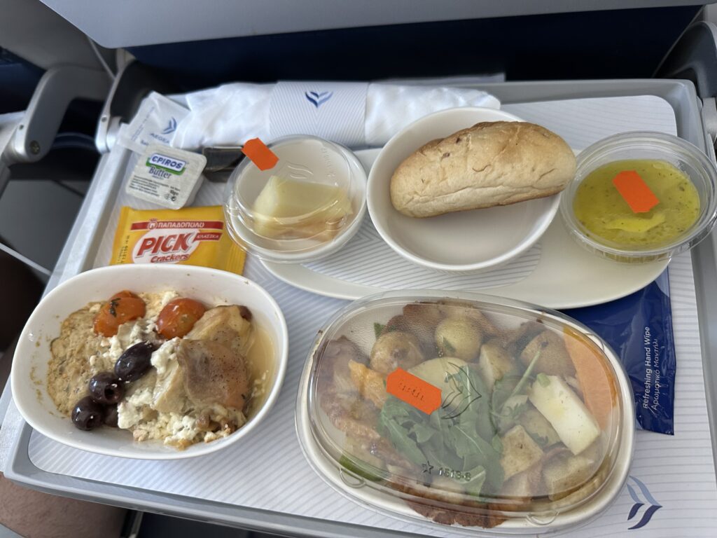 An Aegean meal in Eurobiz is being displayed on the aircraft table. The meal consists of a chicken dish, potato dish, soup and a dinner roll. 