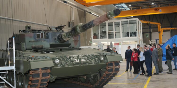 A Leopard 2A4 main battle tank is seen inside a warehouse facility, where 15 onlookers stand by its left side while inspecting it. The tank is painted in camouflage colors.