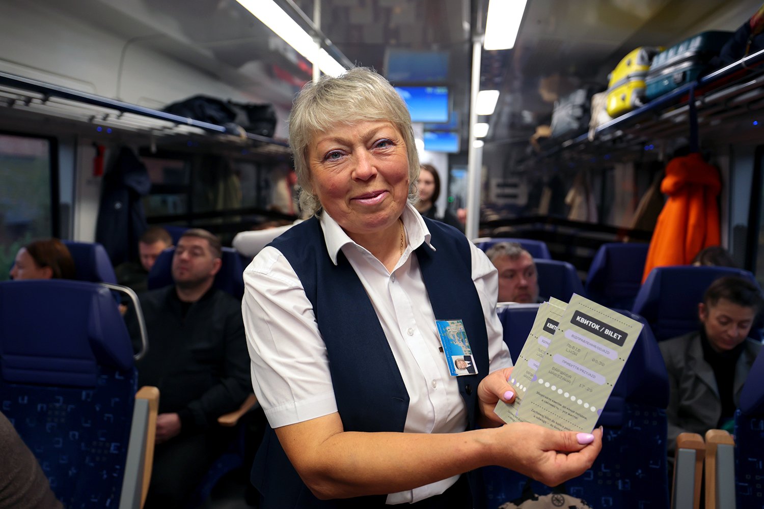 A smiling train guard wearing a blue vest and a white collared shirt displays souvenir tickets on a crowded train, commemoratingthe launch of the international route connecting Lviv and Warsaw.