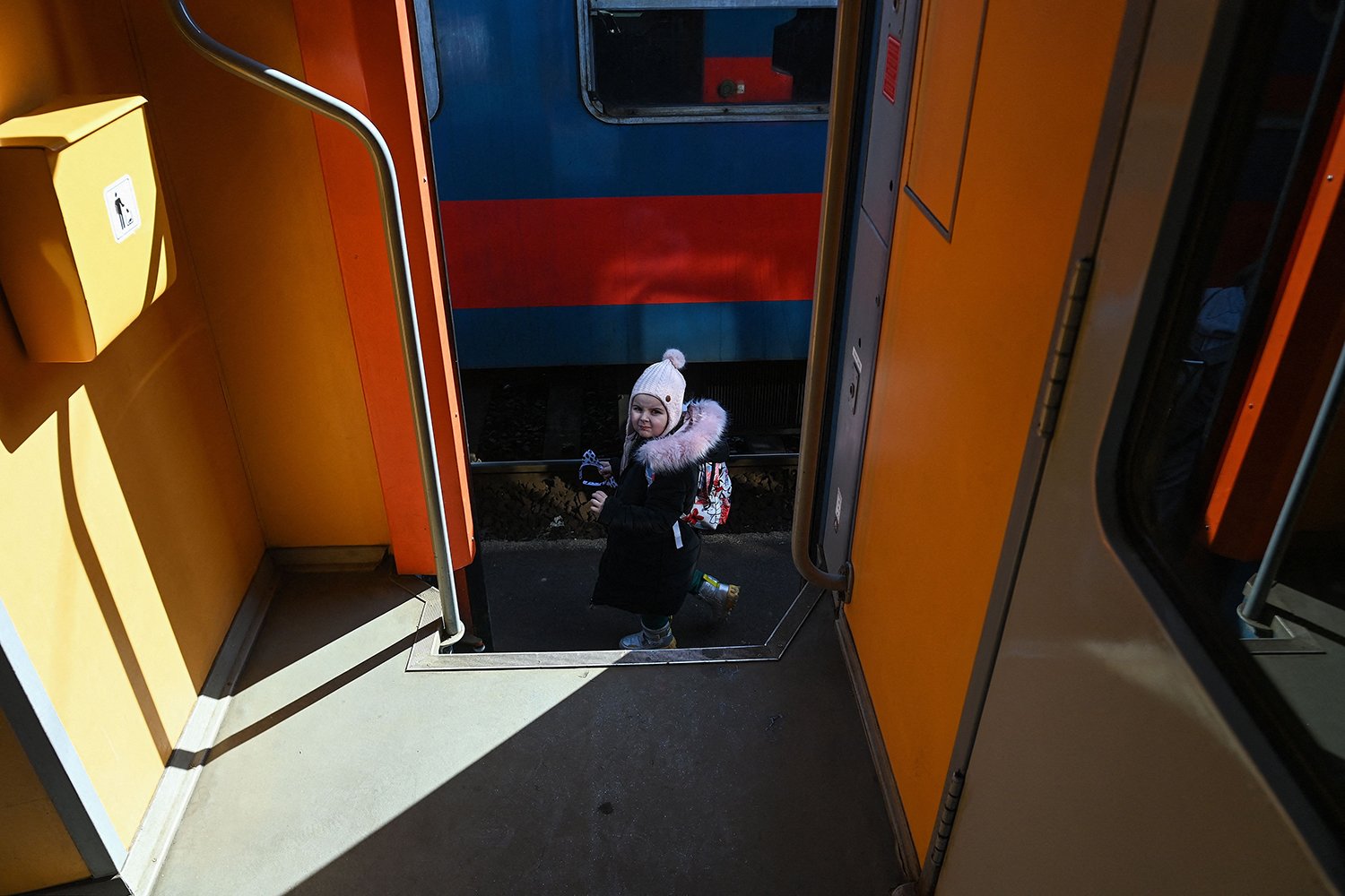 A child, dressed in a white hat and a long black winter coat, walks past the entrance to a train with orange interior walls in the Hungarian-Ukrainian border town of Zahony.