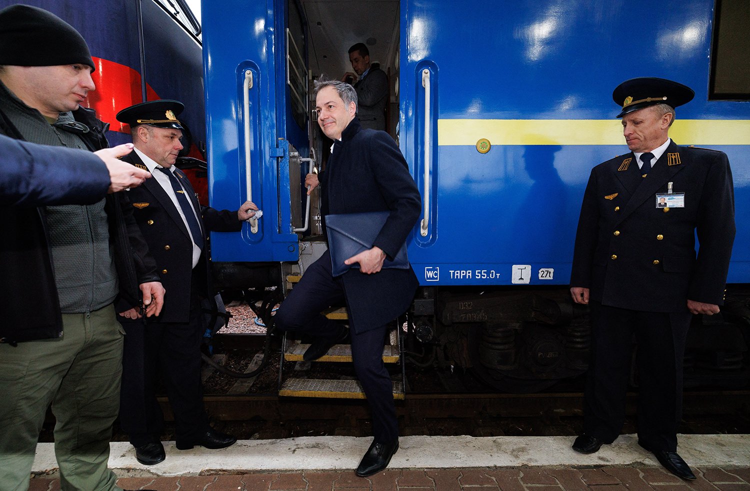 Belgian Prime Minister Alexander De Croo smiles as he steps off a blue train with a yellow stripe in Kyiv, Ukraine, flanked by two men in black uniforms.