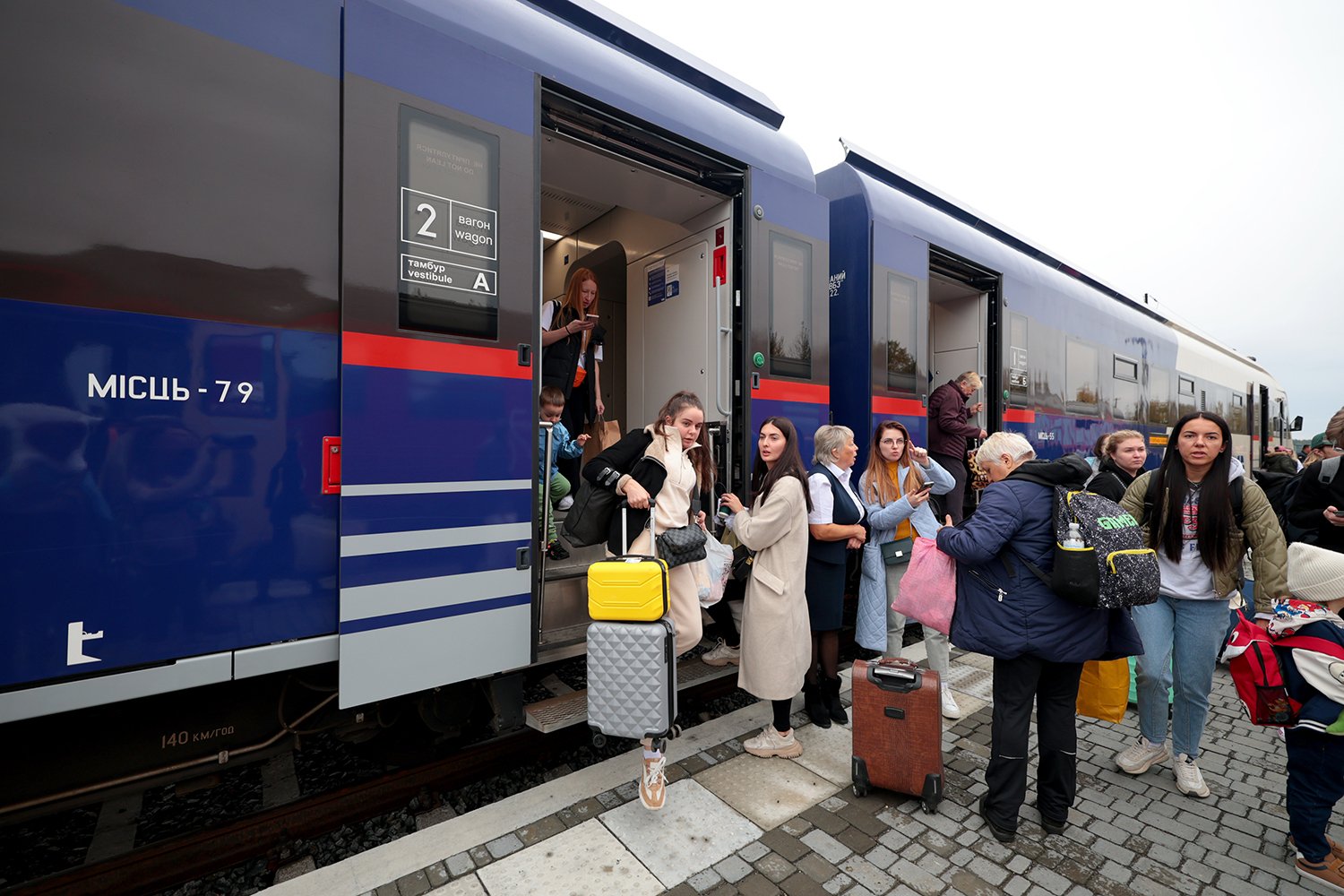 A group of passengers holding luggage and bags enter and exit a blue train with a red stripe and several red stripes as they transfer from a Ukrzaliznytsia train to a Polish SKPL transport company train.