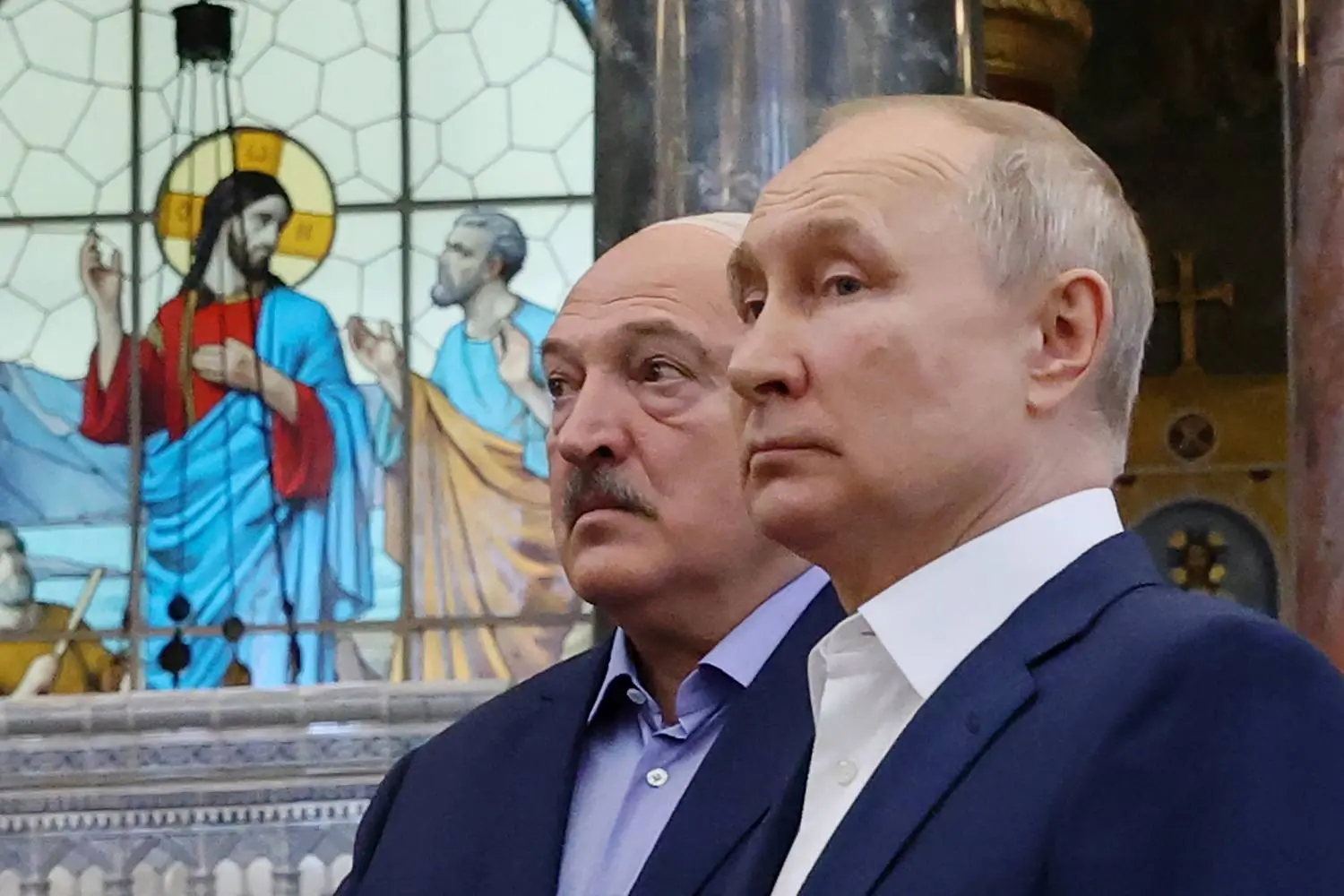 Putin and Lukashenko stand side by side in front of a stained glass window featuring religious imagery.