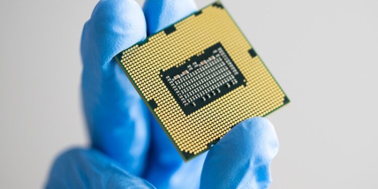 A close-up of a hand wearing a blue protective glove holding a small gold computer chip.