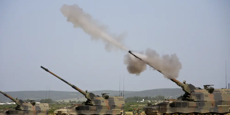 Three tanks, facing left, all have its main gun pointed upwards at a 45-degree angle. The rightmost tank, which is also closest, is in the process of firing ammunition. Smoke is seen shooting out of the gun, with the blurred ammunition in front.