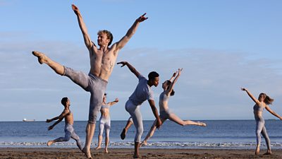 Six professional dancers perform on a sandy beach. Each strikes a different pose. The sea stretches to the horizon in the background.  