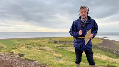 BBC Radio 3 Presenter Petroc Trelawny stands near a coastline, wearing wellington boots and outdoor clothing. He holds a BBC Radio 3 branded microphone and wears headphones. 