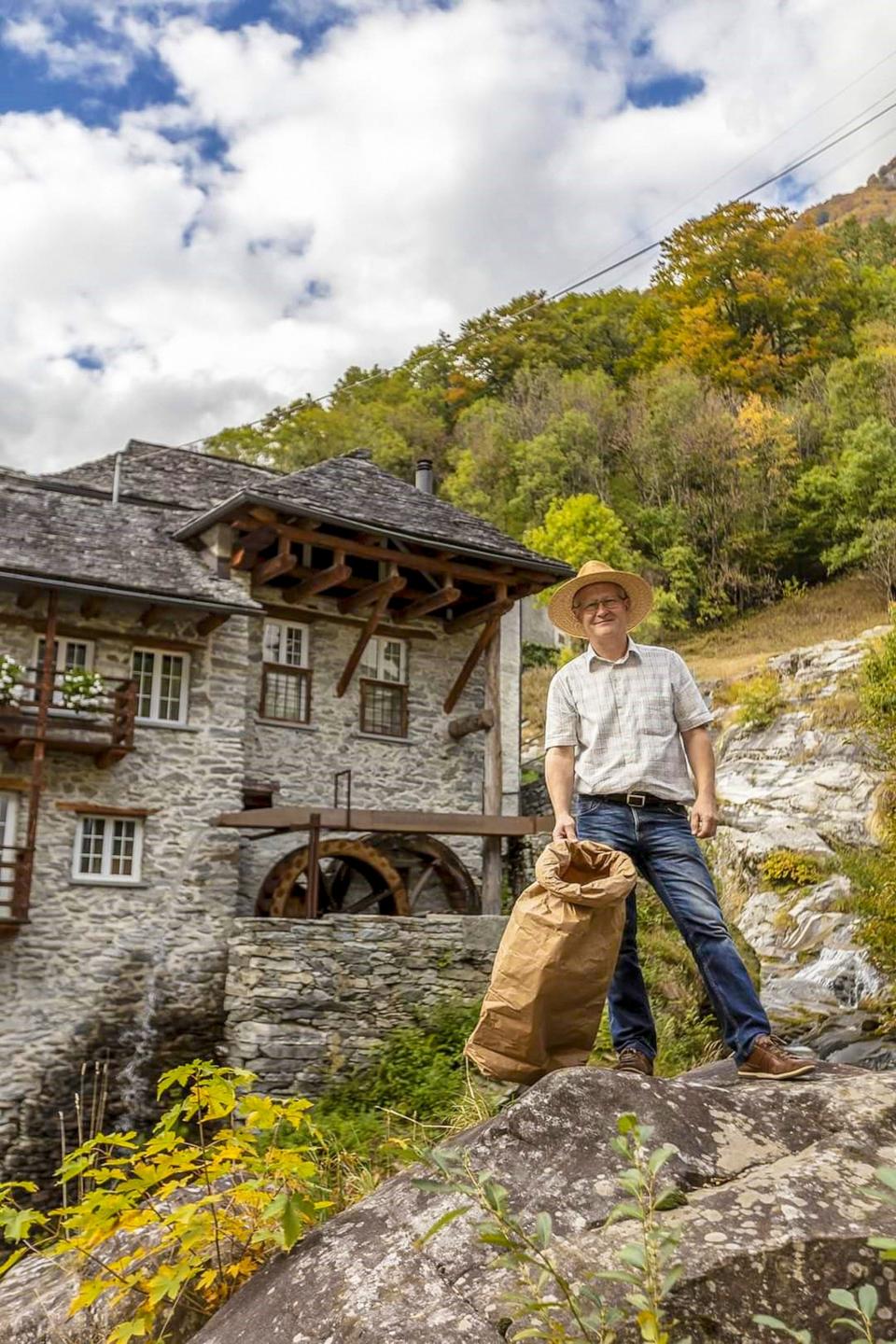 Man stands in front of a stone watermill wearing a straw hat and checked shirt holding a sack