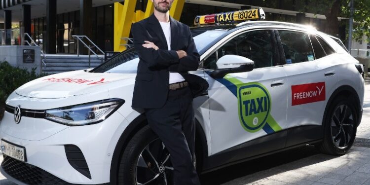 A man wearing a suit, t-shirt and runners stands with arms folded leaning against a white taxi with a FreeNow sticker on the back door.