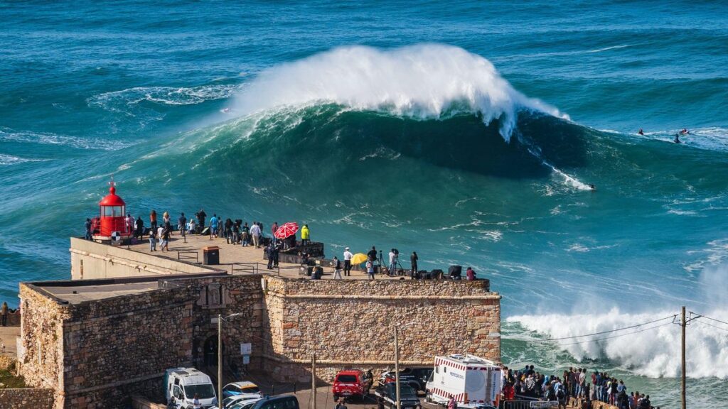 A big wave advances toward the promontory in Nazare, Portugal. There is a small red lighthouse and people watching the wave on the promontory.