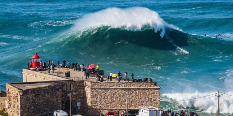 A big wave advances toward the promontory in Nazare, Portugal. There is a small red lighthouse and people watching the wave on the promontory.