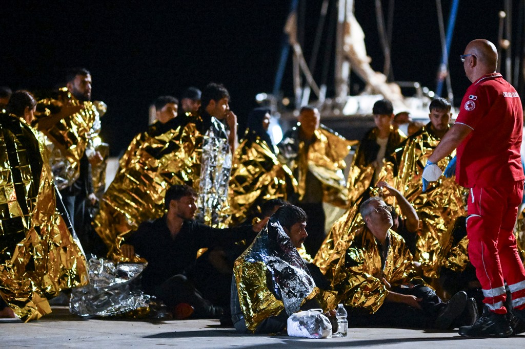 A red cross member in uniform giving instructions to a migrant wearing an aluminum space blanket.