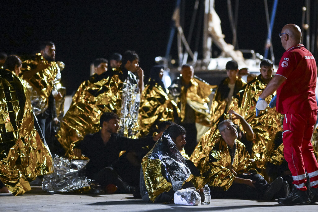 A red cross member in uniform giving instructions to a migrant wearing an aluminum space blanket.