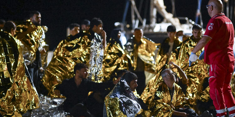 A red cross member in uniform giving instructions to a migrant wearing an aluminum space blanket.