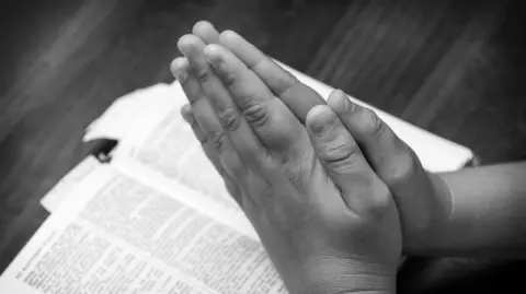 Getty Images A black and white photo showing the hands of a young boy praying over the bible (stock image) 