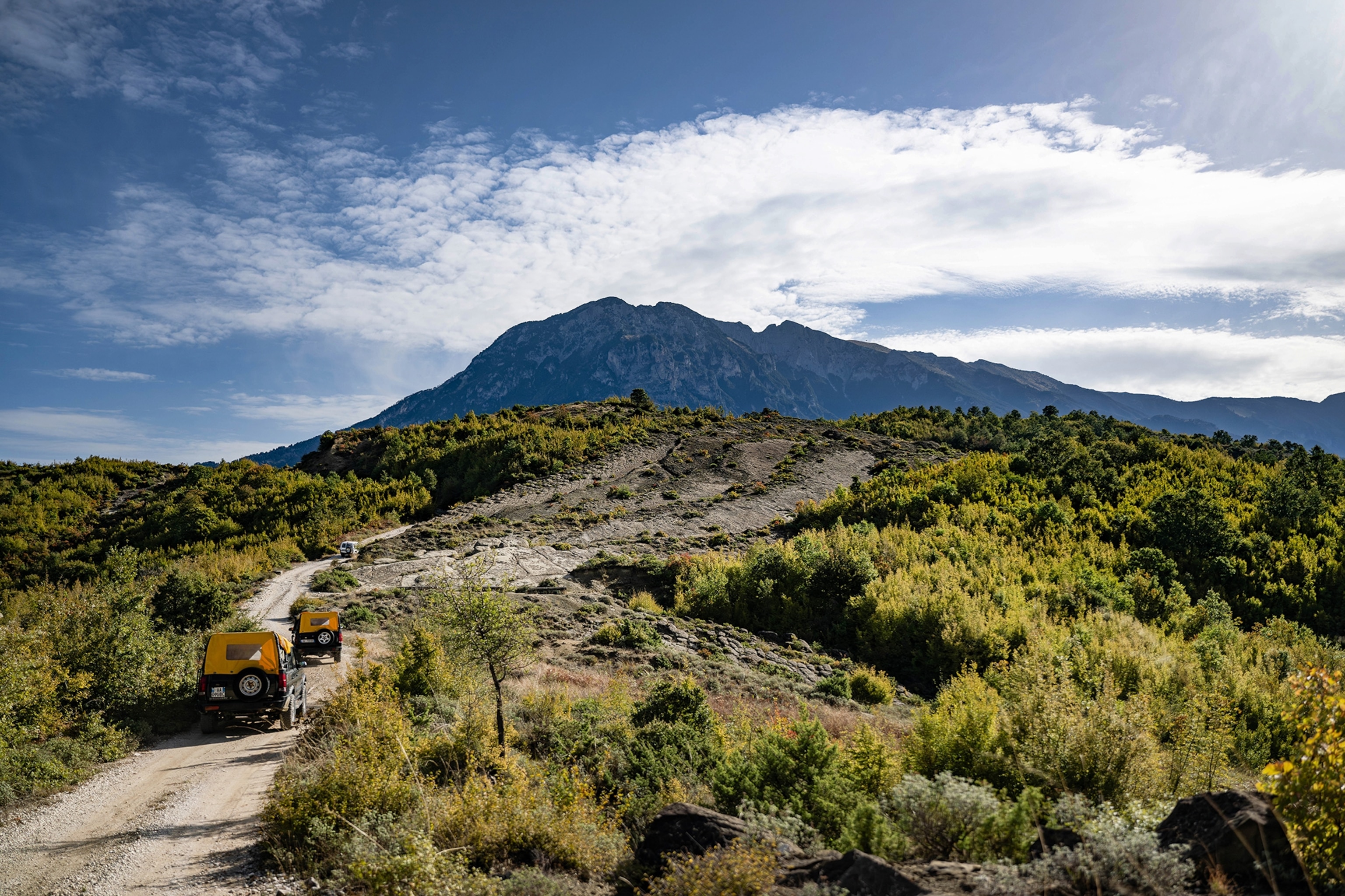 RVs off-roading around the Tomorr Mountain National Park in Albania. A mountain can be seen in the far distance and green hedges line the path.