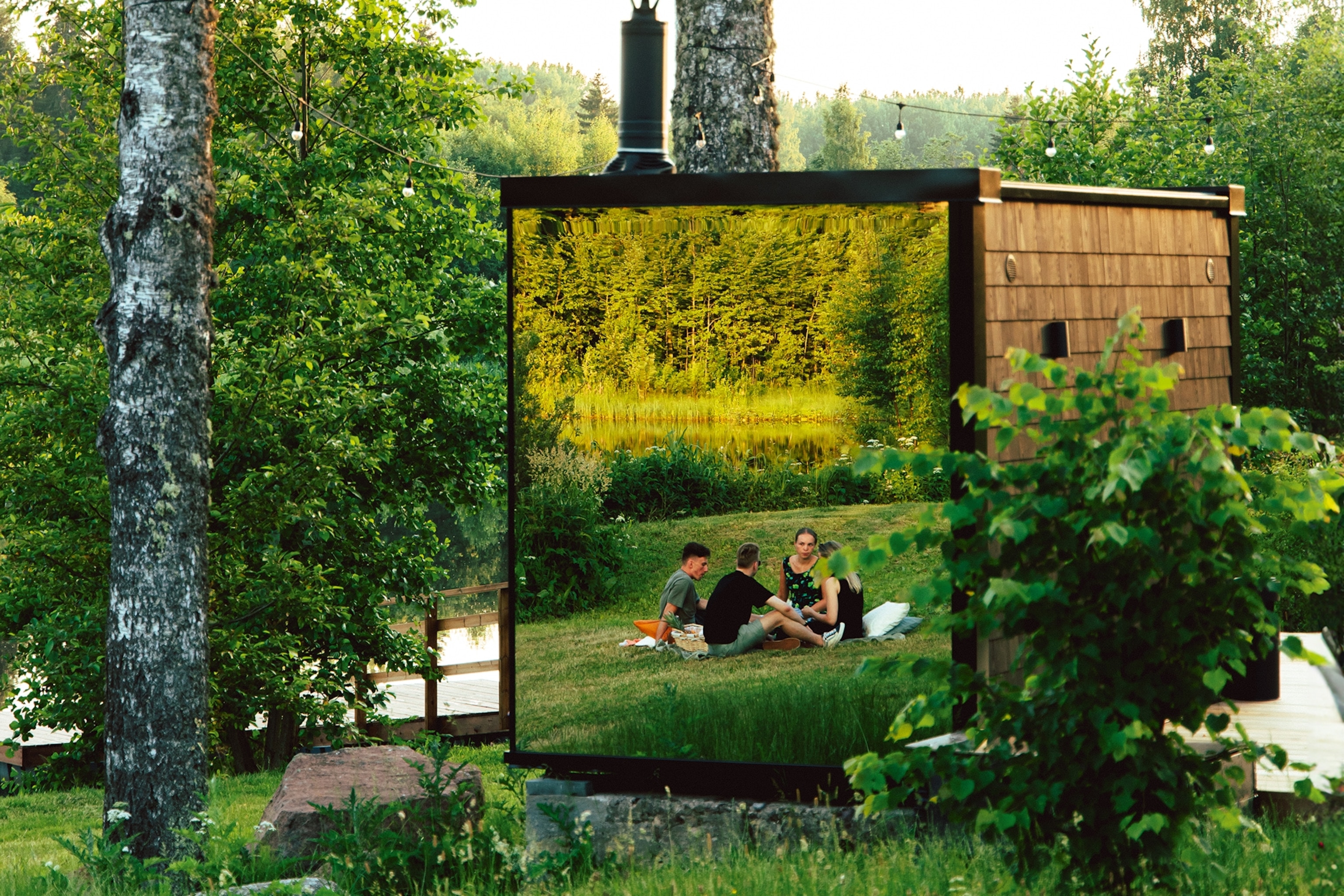 A group of people sitting on grass is reflected on the mirrored surface of a cabin.