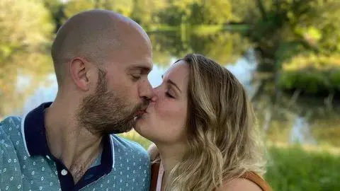 Ainara Pederoso A man with a light beard wearing a light blue patterned polo shirt is to the left of the picture. A woman with shoulder length blonde hair stands beside him. They are kissing.