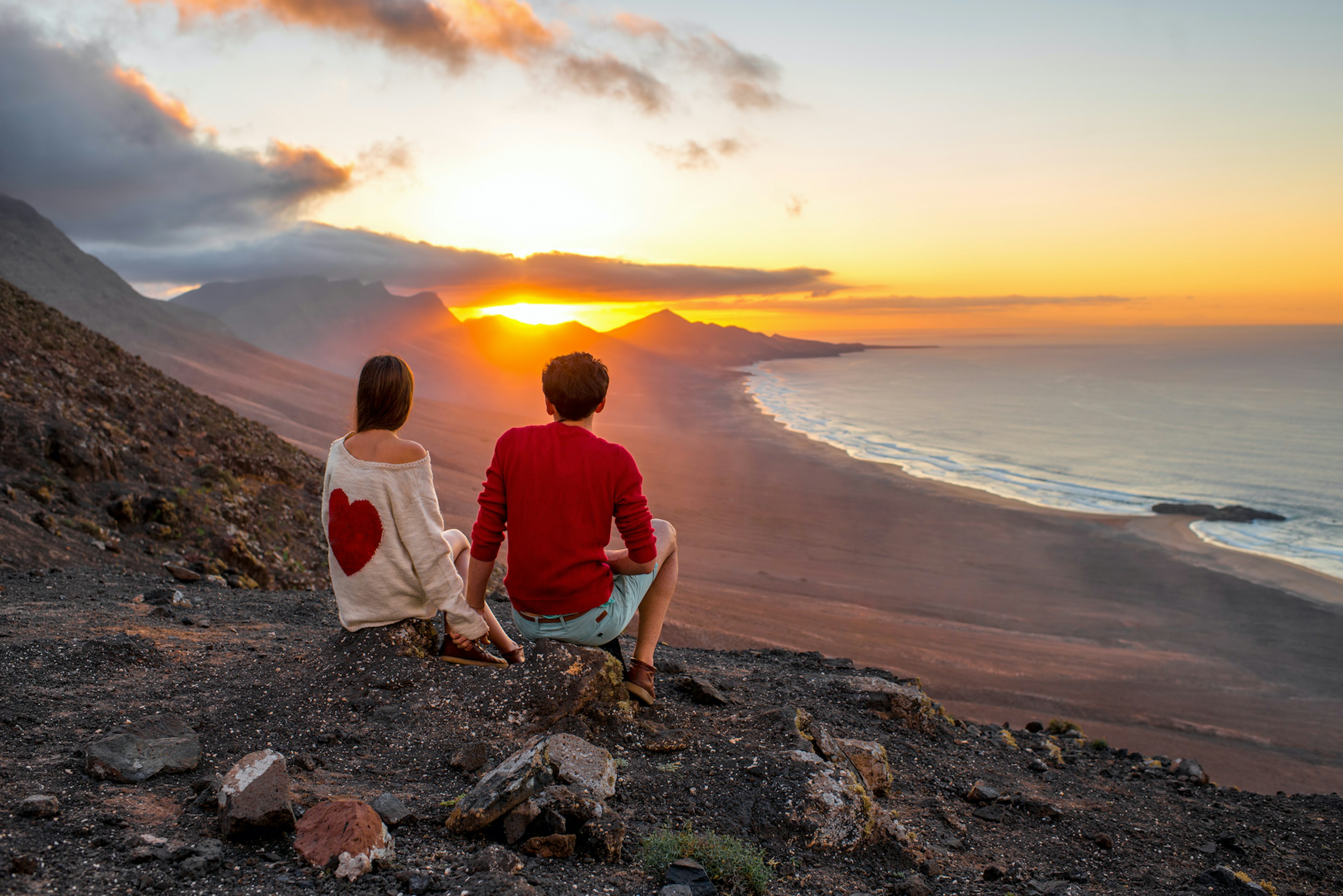 Young couple enjoying beautiful sunset sitting together on the mountain with great view on Cofete coastline on Fuerteventura island.