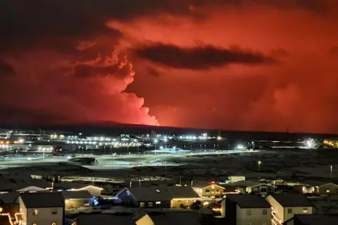 OSKAR GRIMUR KRISTJANSSON Houses in the village of Hafnarfjordur are seen with smoke billowing in the distance, and lava colours the night sky orange - following an volcanic eruption on the Reykjanes peninsula, western Iceland