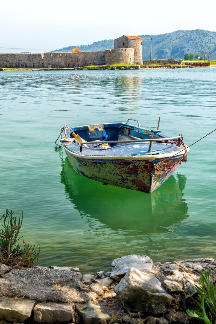 A small boat tethered to shore in blue water, with a harbour wall in the distance