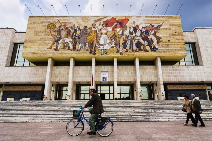 A man cycles in front of a modern building that features a mural of people marching forwards carrying flags, rifles and shields