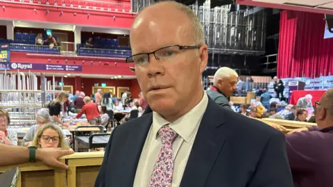 PA Media A man wearing black glasses, a navy suit, white shirt and pink Tie stands in a busy theatre