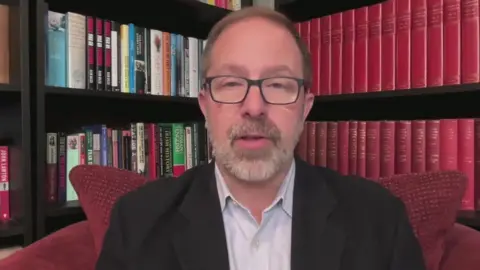 A man in a black suit and white shirt sits in front of a book shelf