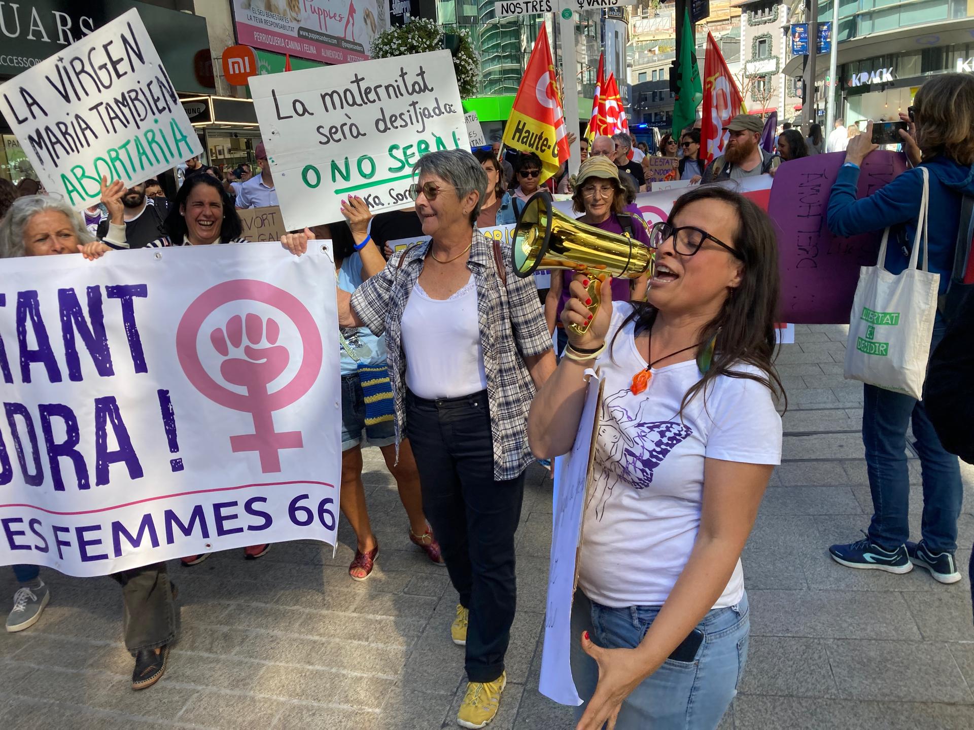 A woman wearing a white shirt and jeans talking into a megaphone with a group of protestors with signs behind her