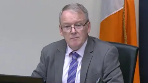 Oireachtas Brian Stanley is sitting in a committee. He's wearing black glasses and a grey suit with a white shirt and a purple tie. The Irish tricolour flag is behind him.