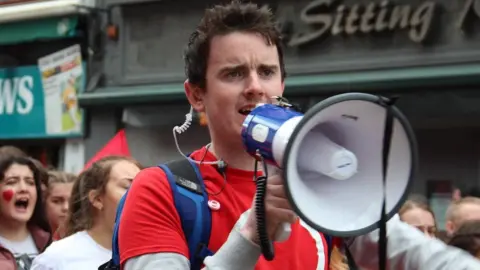 Conradh na Gaeilge Dr Pádraig Ó Tiarnaigh - a man wearing a red shirt holds a megaphone to his mouth while marching in a protest.