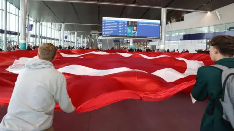 Pacemaker Protestors holding a large red flag with a white circle in the middle stand in the middle of the large main hall of Grand Central Station. The flag is the logo of Irish language group An Dream Dearg