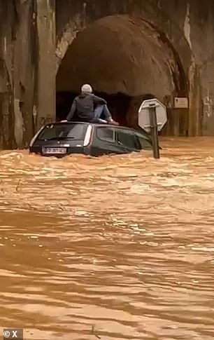 A man is seen sitting a top of a car partially submerged in water