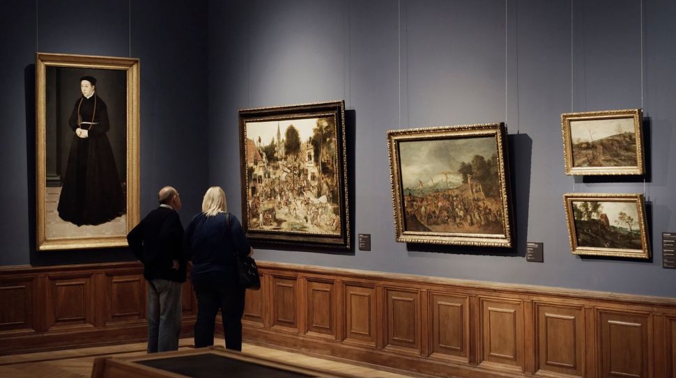 Two people admire large landscape paintings hung on a gray-blue wall.