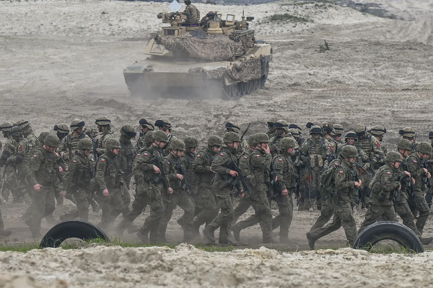 Polish soldiers advancing over a dirt terrain with a tank in the background.
