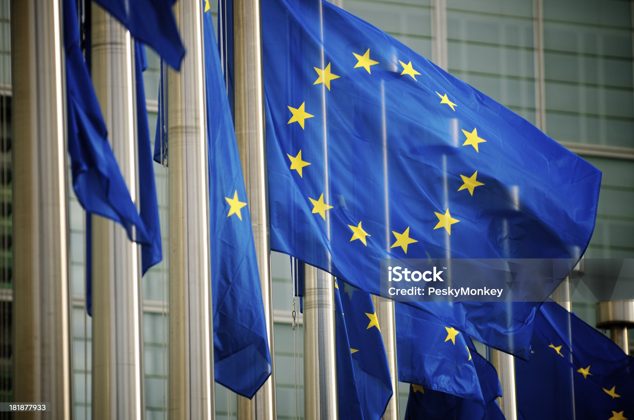 European Union flags waving on poles in front of a modern glass building, partially reflecting sunlight.