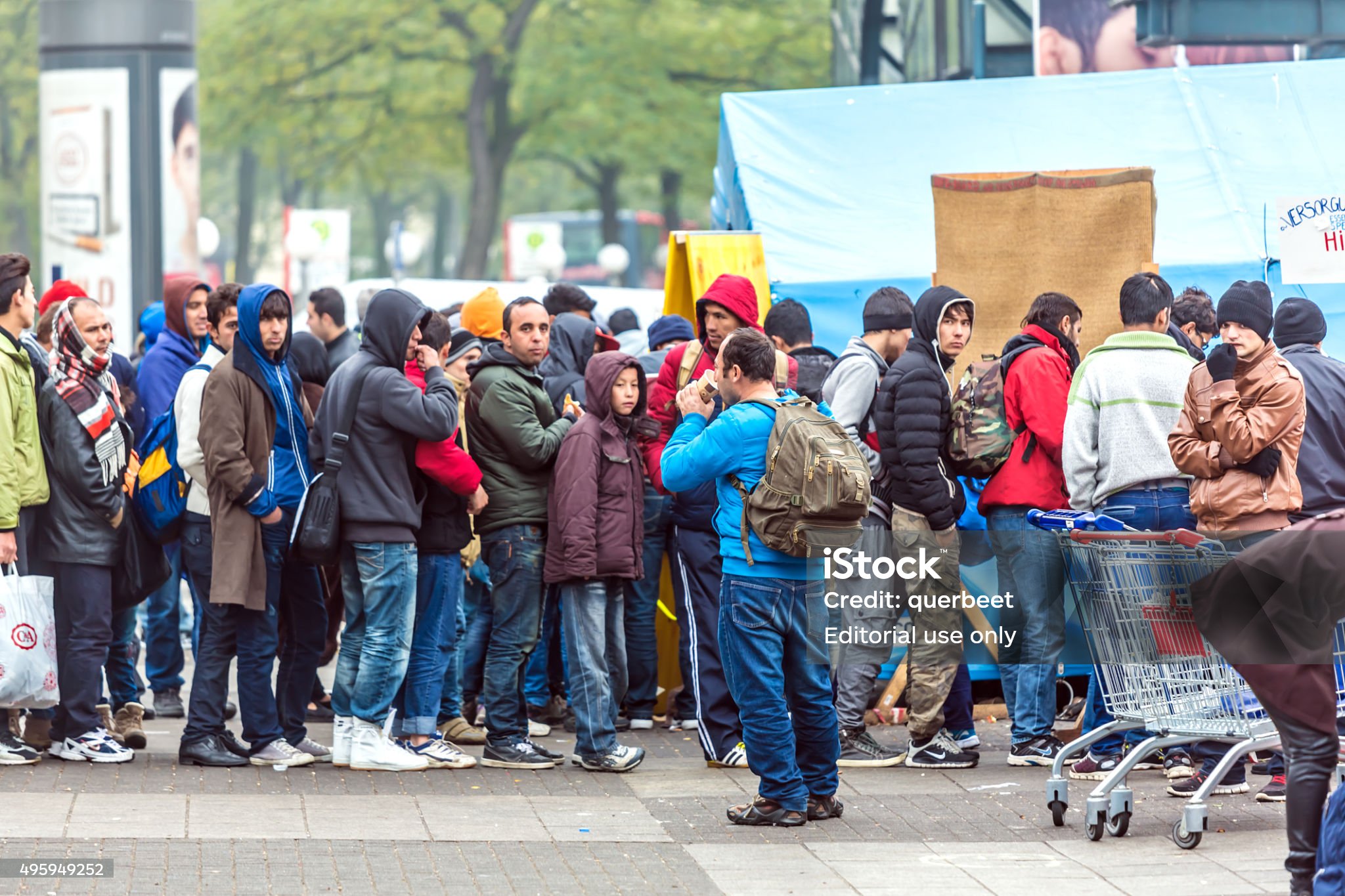 People standing in a long outdoor line, dressed in warm clothes, some holding bags and backpacks.
