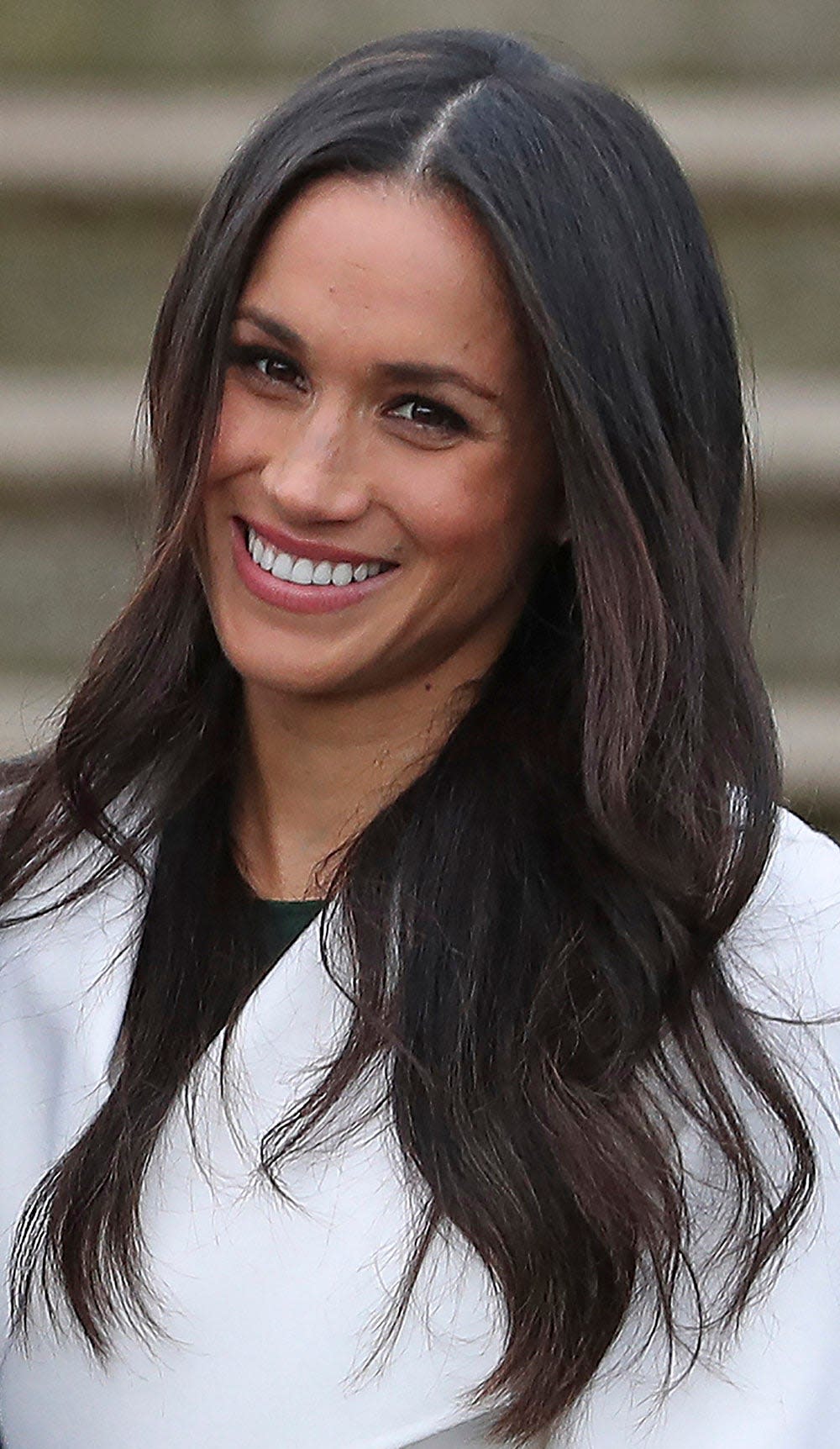 A close-up of Meghan Markle smiling and wearing a white blazer with a black shirt.