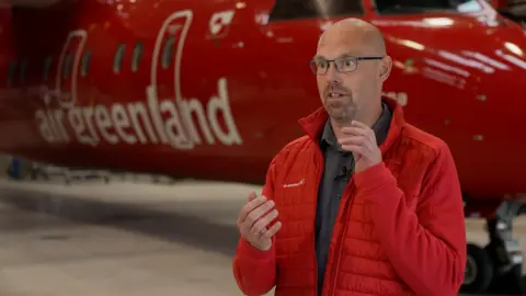 Air Greenland boss Jacob Nitter Sorensen, being interviewed while he stands in front of a plane, while wearing a red jacket.