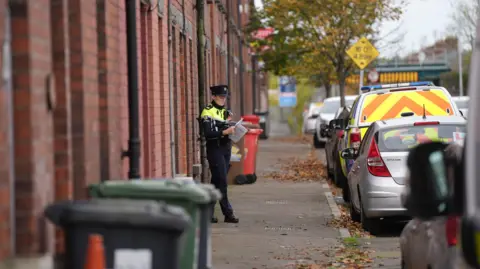 Niall Carson/PA Wire A female garda officer walking out of a house at Emer Terrace, Dundalk. She is wearing full garda uniform and a hat and she is holding documents.