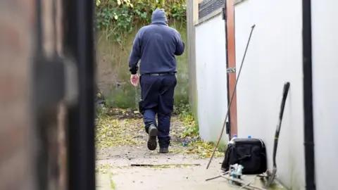 Niall Carson/PA Wire A member of the garda team involved in the search for Kyran Durnin.  The man is wearing dark blue clothing, boots and a beanie hat.   He is walking down an alleyway towards an ivy-covered wall. There is a gardening fork, a bag and bottles in the foreground.