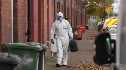 Niall Carson/PA Wire A Gardai forensic officer walking towards the camera on Emer Terrace, a street with red brick terraced houses and lined with parked vehicles. The officer is wearing white hasmat suit and a blue mask and gloves and is carrying a black case. 