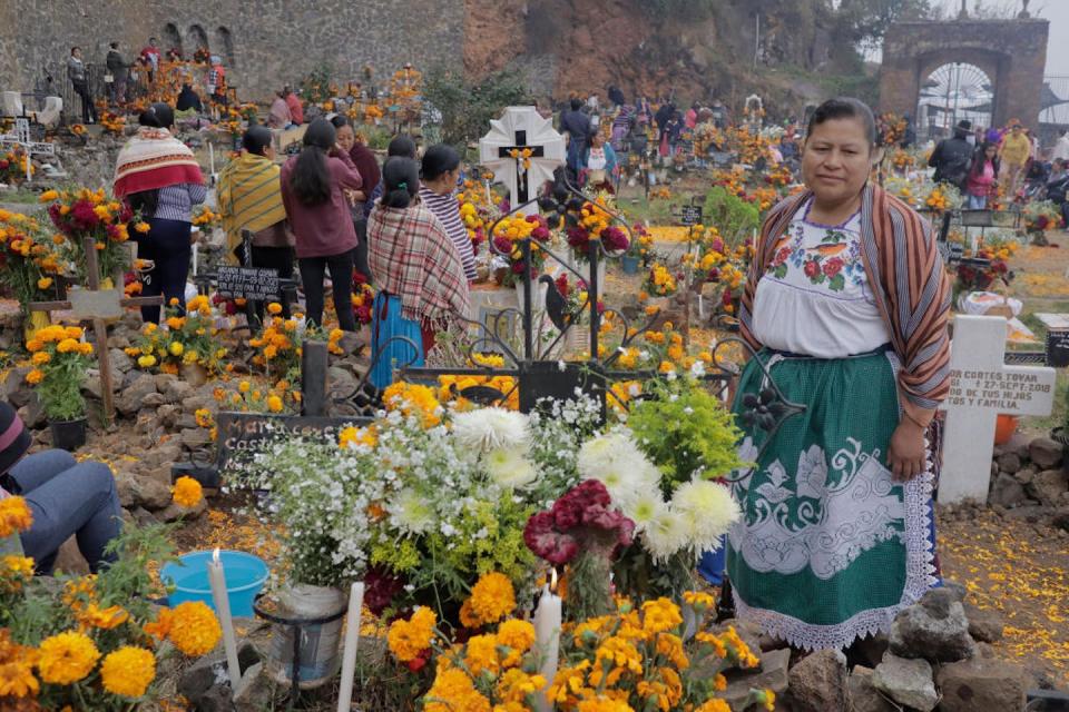 Several people at a gravesite decorated with flowers which has a festive air about it, and one woman looks lovingly at one.