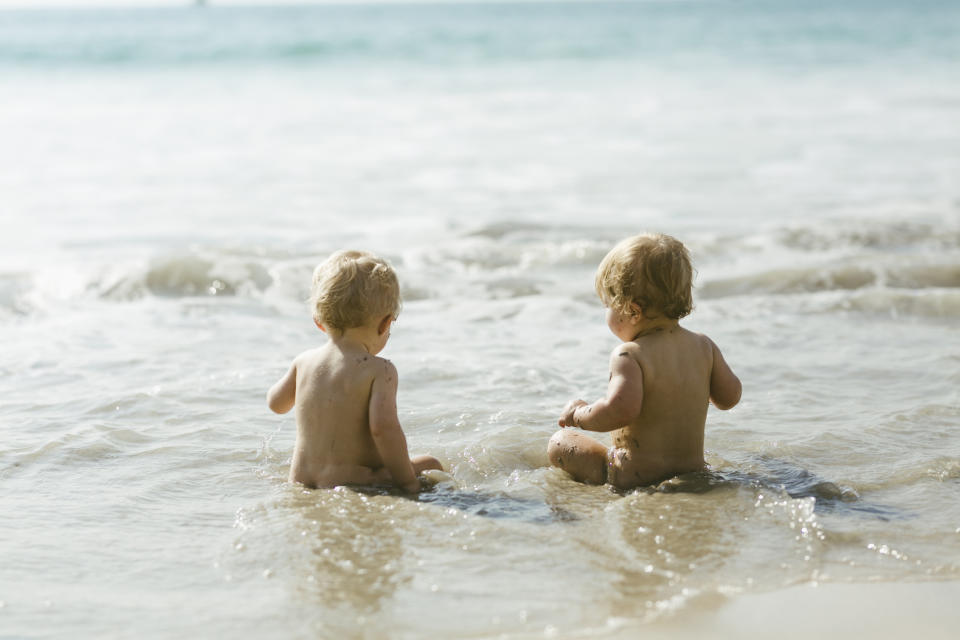 Two toddlers sit at the shore, playing in the shallow water with their backs to the camera. The ocean waves are gently lapping around them