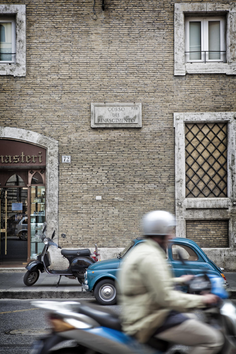 A blurred motorcyclist passes by a parked scooter and small blue car on a street in front of a brick building